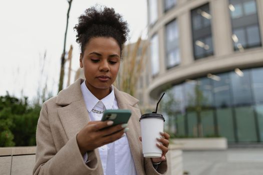 african american young female student looking at a mobile phone with a cup of coffee on the background of the city.