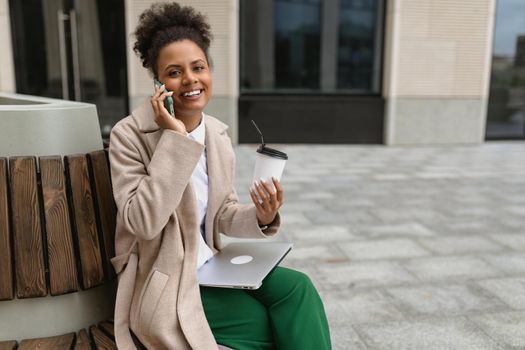 young african american woman sitting near the business center during lunch break with a laptop and a glass of coffee speaks with a smile on the phone.