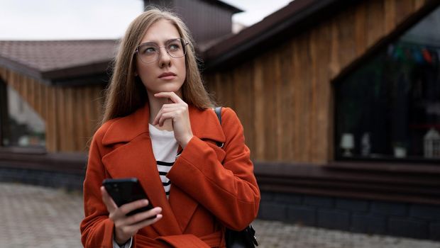 pensive young female student with a mobile phone in her hands against the backdrop of a city building.