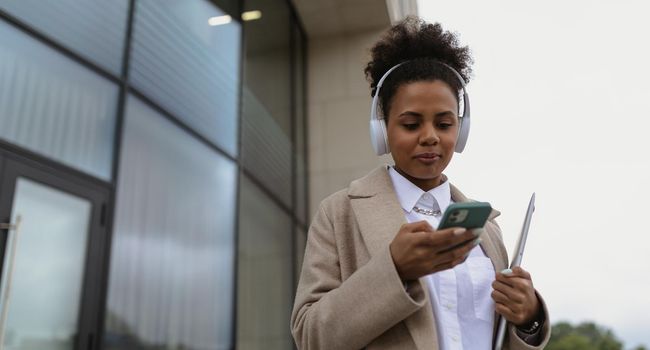 african american young woman in headphones with phone and laptop next to office building.