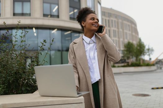 african american woman entrepreneur working on a laptop online and talking on a mobile phone with a smile on the background of a business center.