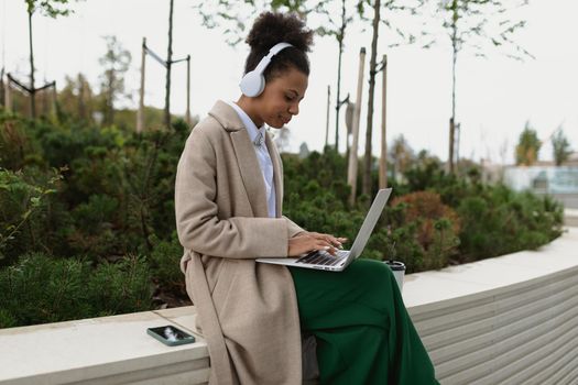 young black female student preparing for an exam working on a laptop online in headphones in a city park.