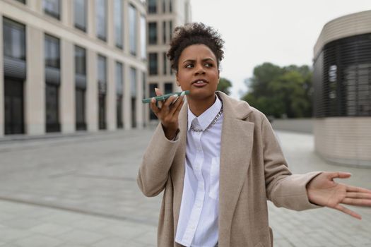 displeased african american woman talking on hands free mobile phone waving her hands against the backdrop of a business center.