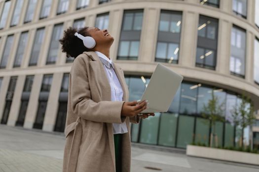 satisfied african american woman with a laptop in her hands and headphones emotionality laughs against the backdrop of an office building.