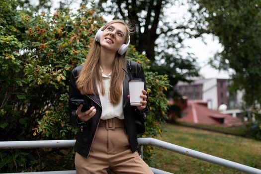 young woman with a cup of coffee by mobile phone listening to music on headphones while relaxing in the park.