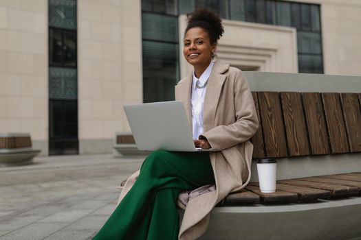 pretty successful young african american woman working on a laptop online sitting on a bench with a glass of coffee on the background of an office building.
