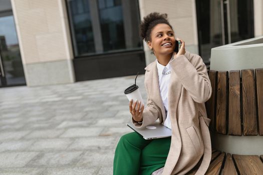 portrait of a young African American female student sitting on a bench with a laptop and talking on a mobile phone against the backdrop of a university in the city.
