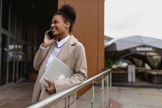 young african american woman talking on a mobile phone with a laptop in her hands next to a business center in the city.