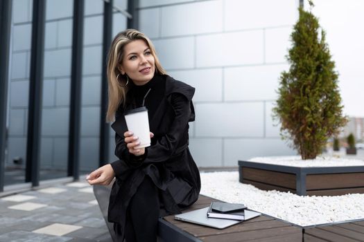 Successful middle-aged businesswoman drinking coffee next to an office building during her lunch break with a smile on her face.