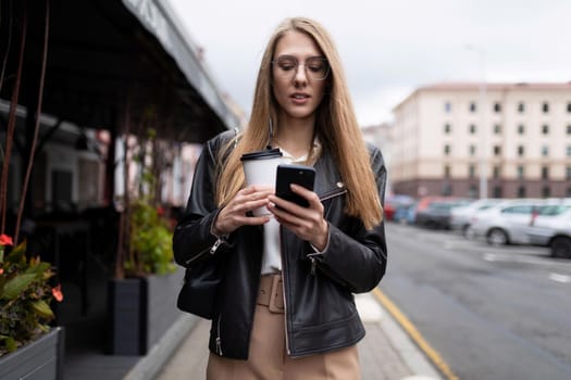 young woman typing a message on a mobile phone with a cup of coffee in her hands against the backdrop of the urban landscape next to a cafe.