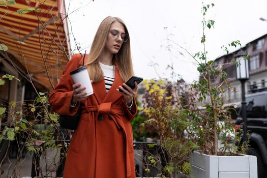 stylish young woman in a warm autumn coat on the background of a city cafe with a glass of hot coffee and a mobile phone in eyeglasses.