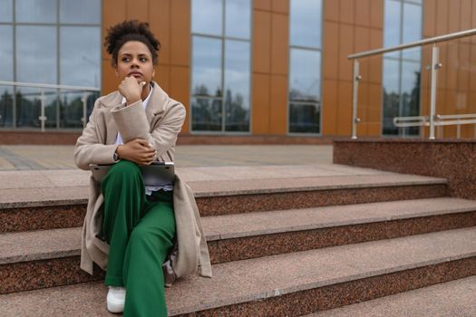 a young African-American woman sits on the steps with a thoughtful face peering into the distance in business clothes against the backdrop of business buildings.