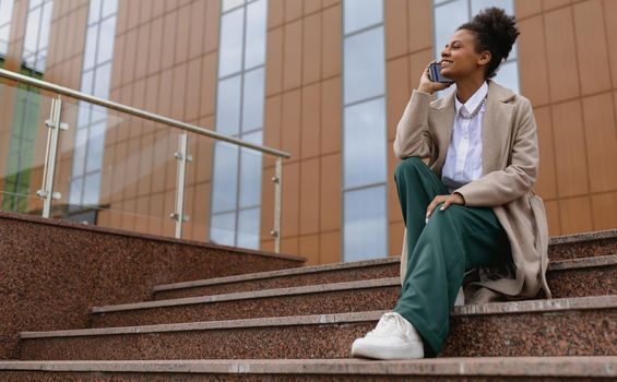 african american young woman talking on the phone while sitting on the steps against the backdrop of a business center.