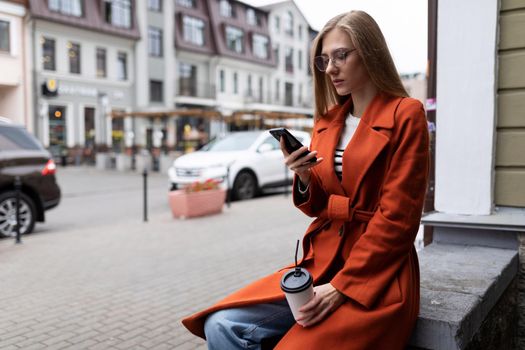 a young student with a cup of coffee looks at a mobile phone on the background of the city.