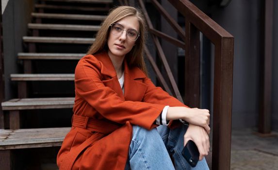 portrait of a pensive young woman in an autumn coat sitting on the stairs next to a cafe with a mobile phone in her hands.