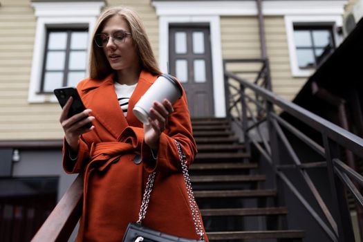 a young woman in an orange coat stands on the stairs with a cup of coffee and a mobile phone.
