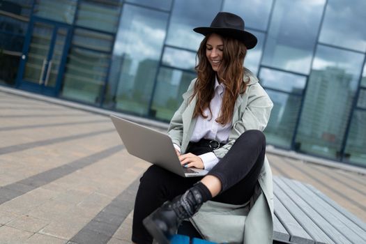 a young woman works on a laptop online against the background of a glass facade of an office building in the city.