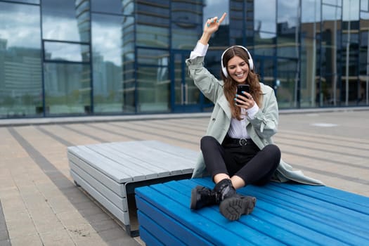 young student listening to music Sitting on a bench in a yoga position dancing against the backdrop of a city building.