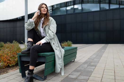 young woman talking on the phone sitting freely on a bench against the backdrop of an office building.