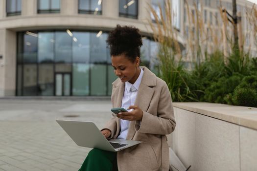 young african american woman student working online on laptops with mobile phone on the background of urban modern building.