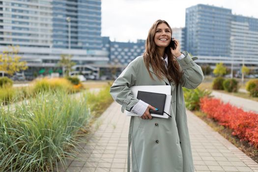 young woman student with a laptop weekly under her arm talking on the phone on the background of the city.
