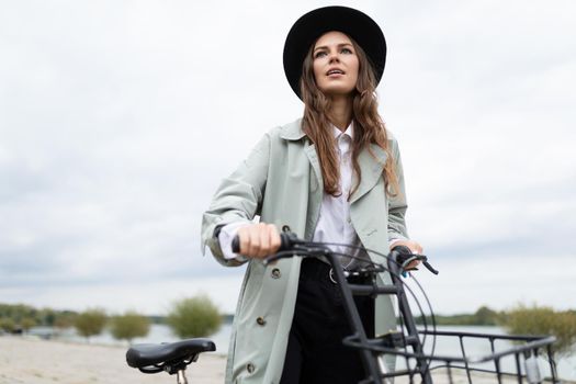 young stylish woman in a raincoat and hat rides a bicycle along the promenade.