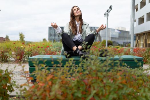 a young woman sits on a bench in the park in a yoga pose with her arms apart with her eyes closed.