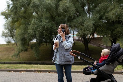 a young mother with a baby in a stroller walks in the park with a cup of coffee in her hand and speaks on a mobile phone.