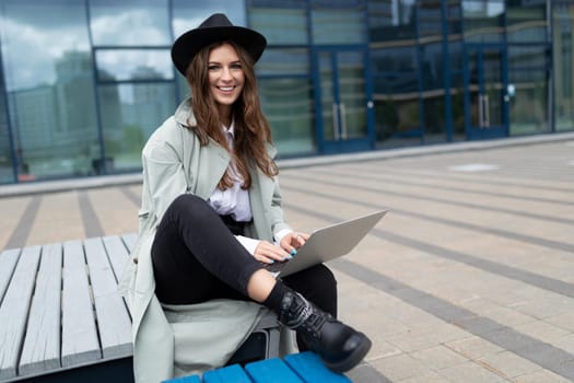 young stylish woman sitting on a bench with a laptop in a hat smiling mischievously looking at the camera.