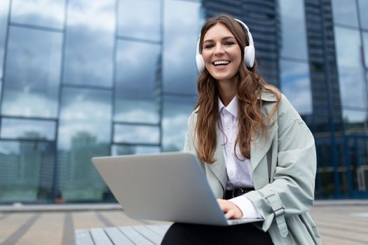 a young student sits on a bench with a laptop in headphones and smiles looking at the camera against the backdrop of a glass building.