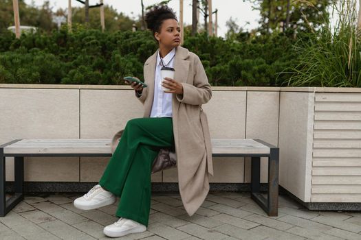 an african american woman sits on a bench in the park with a laptop and mobile phone looks thoughtfully into the distance.