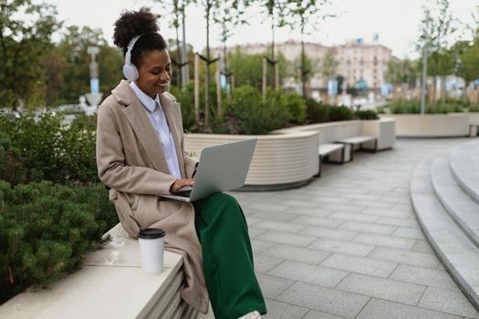 african american young woman working online on a laptop in headphones with a cup of coffee on the background of the city with a smile on her face.