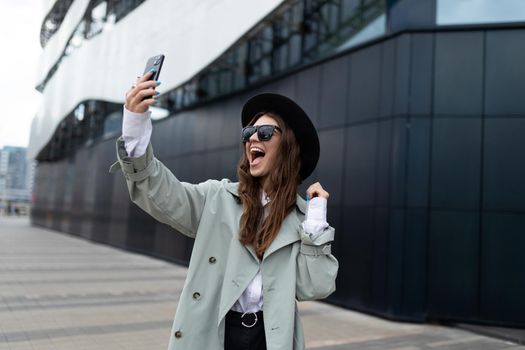 a strong young woman takes a selfie against the backdrop of an office building in a black hat and autumn gray coat looking at the phone camera with a smile.