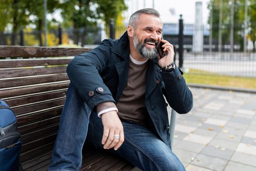 a mature man with a gray beard is talking on the phone with a smile while sitting on a bench in a city park.