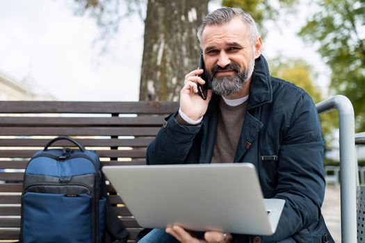 an elderly business man speaks on a mobile phone while working on a laptop online in a park on a bench.
