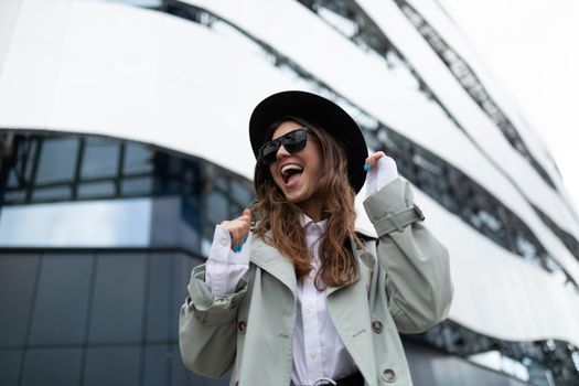 successful young emotional woman dancing and grimacing with a smile on her face against the backdrop of a modern stylish building.