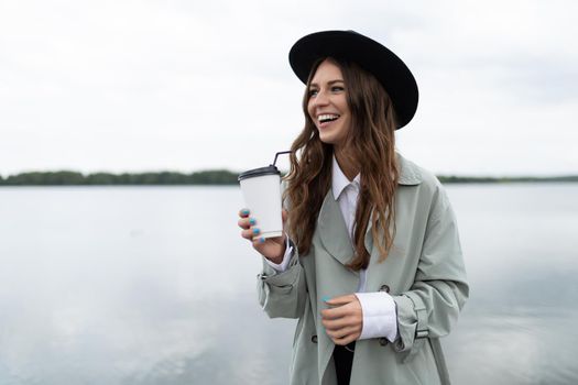 a young woman walks along the embankment against the backdrop of a large lake with a glass of coffee with a smile and in a black hat.