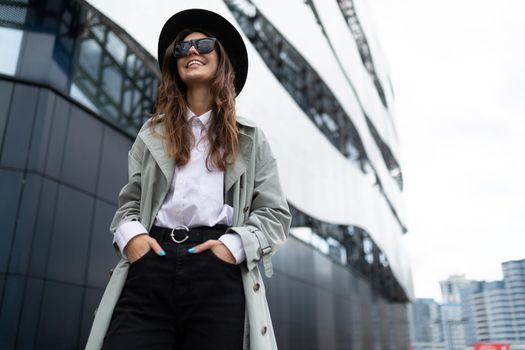 young stylish woman in sunglasses in a black hat walks around the city against the backdrop of office buildings with a smile on her face.