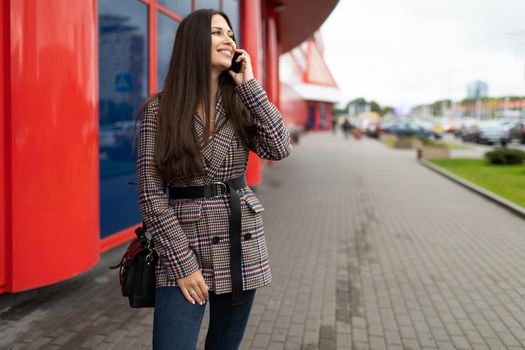 Beautiful stylish young woman talking on a mobile phone with a smile on her face against the background of a red blue city building facade.