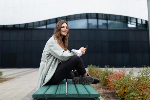 a young woman sits on a bench cross-legged under her with a mobile phone looking at the camera on the background of the city.