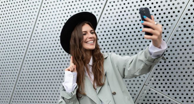 stylish young woman in a black hat and gray coat takes a selfie against the backdrop of a non-aluminum facade of an office building.