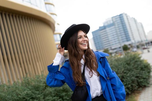 portrait of a strong young woman in a black hat and blue raincoat walking around the city with a smile on her face.
