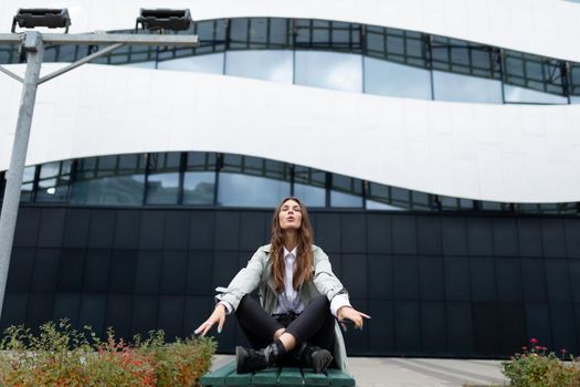 a young woman sits in a yoga position against the backdrop of an office building on a bench with her eyes closed, arms spread apart.
