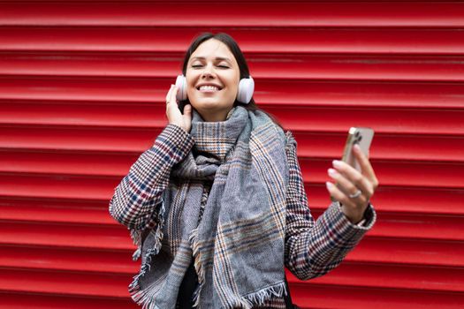young and happy woman in headphones closing her eyes listens to music with a wide smile on her face through a mobile phone against the background of a red bright wall.