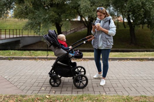 a young mother drinks coffee and watches a mobile phone while walking with a baby in a stroller around the city.