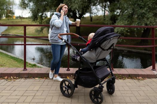 young caring mother with a cup of coffee in her hands next to the stroller with her newborn child while walking in the park speaks on the phone with a smile on her face.