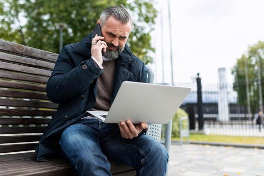 on the way to the office, a man is talking on the phone and looking at a laptop while sitting on a bench next to the business center.
