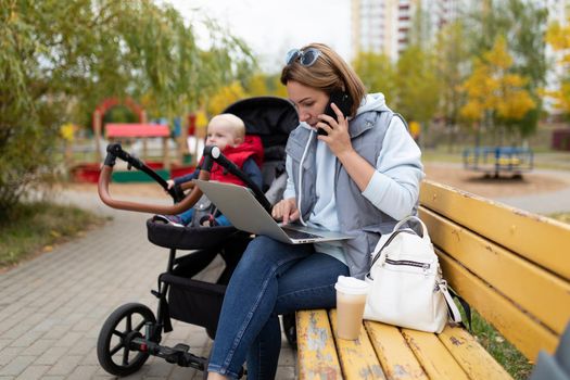 a young mother sitting on a bench next to a stroller with a child works online on a laptop and answers a phone call.