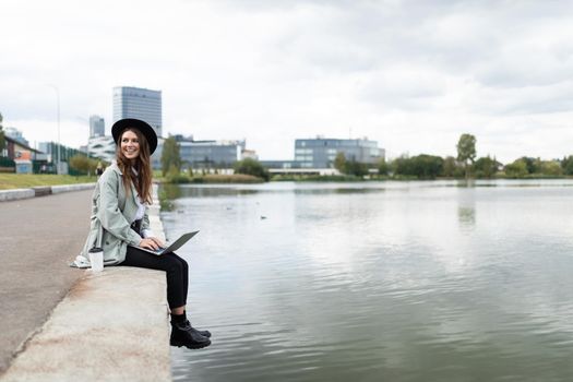 strong young woman working online on a laptop sitting on the promenade dangling her legs in the water with a smile on her face.