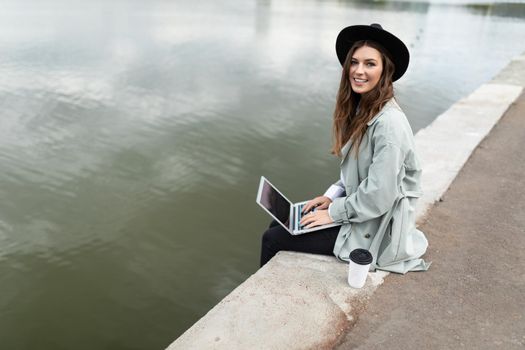 young woman working on a laptop online sitting on the promenade dangling her legs to the water with a smile looking at the camera.
